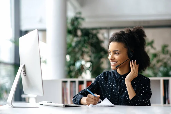 Afro-américaine amicale jeune femme avec casque, travailleur de centre d'appels, consultant, homme d'affaires, regarde le moniteur d'ordinateur tout en communiquant avec des collègues ou des clients, effectue des consultations — Photo