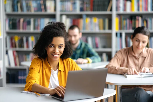 Studenten sitzen während einer Vorlesung an ihren Schreibtischen in der Bibliothek. Afroamerikanisch gelockte hübsche Studentin sitzt während einer Unterrichtsstunde mit Laptop an einem Tisch, blickt freundlich lächelnd in die Kamera — Stockfoto