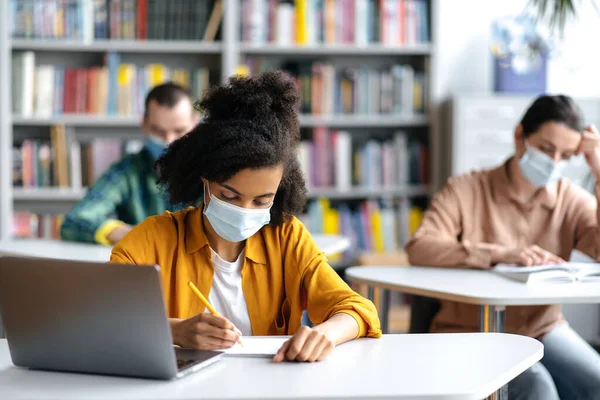 Aprendendo durante uma pandemia. Estudantes em máscaras médicas protetoras se sentam na biblioteca da universidade a uma distância um do outro. Estudante afro-americana toma notas durante palestra — Fotografia de Stock