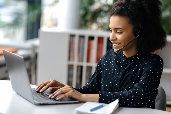 Wanita muda Afrika Amerika yang cantik mengenakan headset, duduk di meja kerja, menggunakan laptop, bekerja online, berselancar di Internet, mengobrol dengan rekan atau klien, menjawab email, tersenyum — Stok Foto