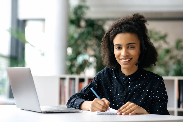 Charmante jeune femme afro-américaine à succès, dans un casque, pigiste, employée ou gestionnaire, assise à une table de travail avec un ordinateur portable et un carnet, prend des notes, regarde la caméra, sourit amical — Photo