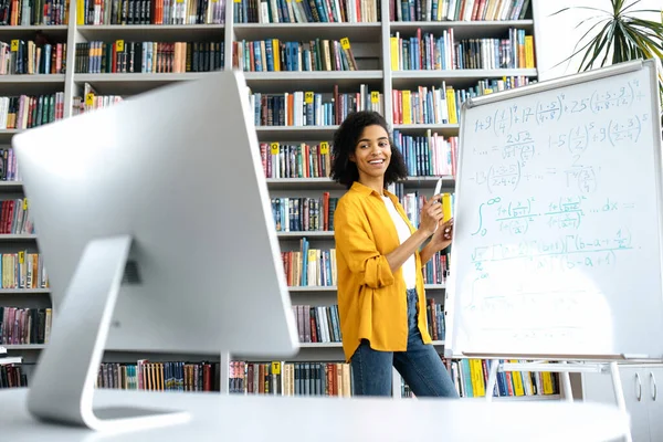 Ευχάριστη αφροαμερικανίδα δασκάλα διεξάγει online μάθημα για τους μαθητές μέσω τηλεδιάσκεψης από υπολογιστή, στέκεται στη βιβλιοθήκη του πανεπιστημίου, δείχνει πληροφορίες στο λευκό πίνακα, κοιτάζει τον υπολογιστή, χαμόγελο — Φωτογραφία Αρχείου
