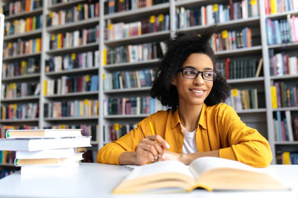 Adolescente afroamericana bella studentessa con gli occhiali, che studia mentre siede a tavola in una biblioteca universitaria, legge libri alla ricerca di informazioni per una lezione o un esame, facendo i compiti, distoglie lo sguardo — Foto Stock