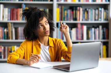 Oldukça kıvırcık saçlı zeki Afro-Amerikalı kız öğrenci elinde dizüstü bilgisayarla kütüphanede oturuyor, düşünceli bir şekilde bir öğretmenden ödevini bitirmeyi düşünüyor.