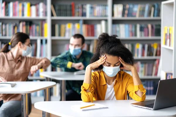 Uma estudante afro-americana cansada com uma máscara médica protetora senta-se em uma mesa em uma biblioteca com um laptop, olhos fechados, pensa. No fundo, dois alunos cumprimentam-se com um galo de cotovelo — Fotografia de Stock