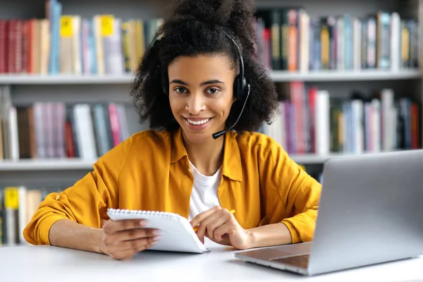 Étudiante afro-américaine avec un casque, assise à une table dans la bibliothèque, tenant un cahier dans sa main, utilisant un ordinateur portable pour l'enseignement à distance, regardant la caméra, souriant — Photo