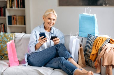 İnternetten alışveriş. Beyaz tenli, şık, orta yaşlı kadın, oturma odasında koltukta oturuyor, akıllı telefon ve kredi kartını internet üzerinden ödeme için kullanıyor, internetten sipariş veriyor, gülümsüyor.