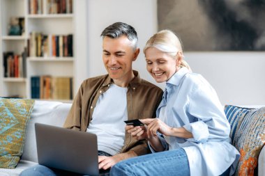 İnternetten alışveriş. Başarılı Kafkasyalı, aşık olgun bir çift, rahat giysiler içinde kanepede oturan mutlu karı koca, bilgisayar ve kredi kartı ile mal veya teslimatı yapan, gülümseyen