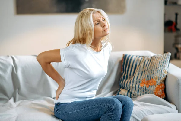 Back pain in a mature woman. Unhappy frustrated middle-aged caucasian woman, upset blonde sitting on the sofa in the living room, feeling joint problems, needing rest, looking away, sad
