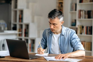 Odaklanmış, gri saçlı yönetici, koç ya da serbest çalışan, şık kıyafetler giyen, evde dizüstü bilgisayar ve belgelerle çalışan, bir iş projesi hazırlayan, notlar alan
