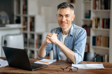 Kendine güvenen, beyaz saçlı, olgun beyaz saçlı, borsacı ya da yönetici portresi, şık günlük giysiler içinde, evde dizüstü bilgisayarda çalışıyor, çalışma masasında oturuyor, kameraya bakıyor, dostça gülümsüyor.