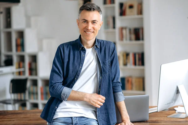 Portrait of a handsome confident successful mature gray-haired caucasian man, in casual stylish denim shirt, stands at the work desk, looks at the camera, smiles friendly