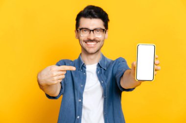 Gözlüklü beyaz tenli adam kameraya bakıyor, sunumunuz için beyaz ekranlı akıllı telefonu tutuyor ve onu işaret ediyor, izole edilmiş turuncu arka planda duruyor, boşluğu kopyalıyor.