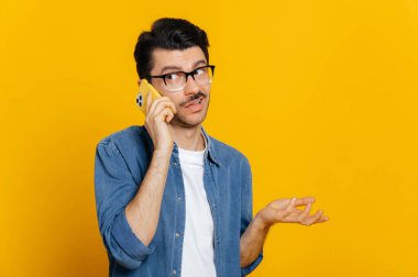 Kafası karışmış, kafası karışmış, gözlüklü, akıllı, hoş olmayan bir şekilde konuşan, öfkeyle başka tarafa bakan ve el kol hareketi yapan, izole edilmiş turuncu bir arka planda duruyor.