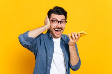 Gözlüklü duygusal beyaz adam, elinde akıllı telefon, telefon görüşmesi sırasında çığlık atıyor ya da sesli mesaj kaydediyor, izole edilmiş turuncu arka planda duruyor, elini başının yanında tutuyor. Ses iletisi