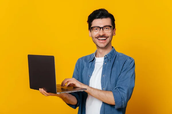 Atraktivní spokojený stylový neoholený inteligentní běloch s brýlemi v ruce, s otevřeným laptopem, pohledy a přátelským úsměvem na kameru, zatímco stojí proti izolované oranžové — Stock fotografie