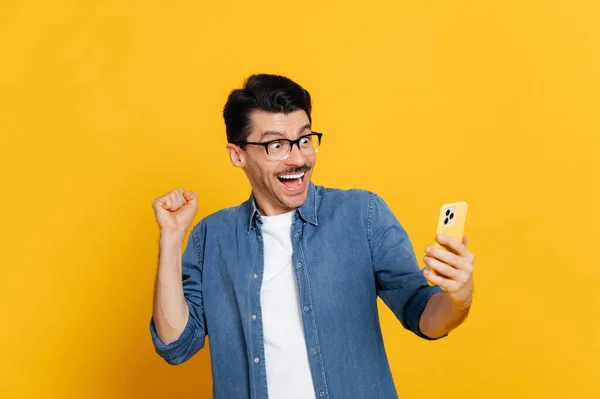 Verbazingwekkende vrolijke opgewonden stijlvolle blanke man houdt smartphone, onverwacht nieuws te krijgen, te winnen, staat op geïsoleerde oranje achtergrond, vrolijke gezichtsuitdrukking, tanden glimlach, gebaren met vuist — Stockfoto