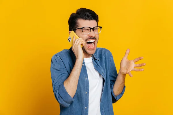 Onaangenaam telefoongesprek. Geïrriteerde boze blanke jongeman schreeuwt tijdens telefoongesprek, gebaart met de hand, staat op een geïsoleerde oranje achtergrond — Stockfoto