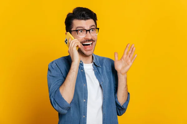Telefoongesprek per mobiele telefoon. Happy vrolijke opgewonden Kaukasische charismatische stijlvolle man heeft een aangenaam telefoongesprek, gelukkige gezichtsuitdrukking, staat op geïsoleerde oranje achtergrond, gebaren hand — Stockfoto