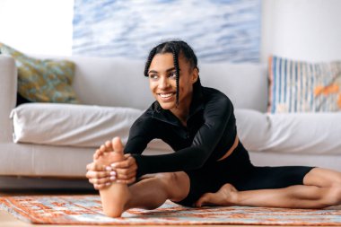 Esnek, aktif, spor kıyafetleri içinde spor giyinmiş, sağlıklı bir hayat sürüyor, evde yere uzanıyor, kollarını ayaklarına dayayıp gülümsüyor.