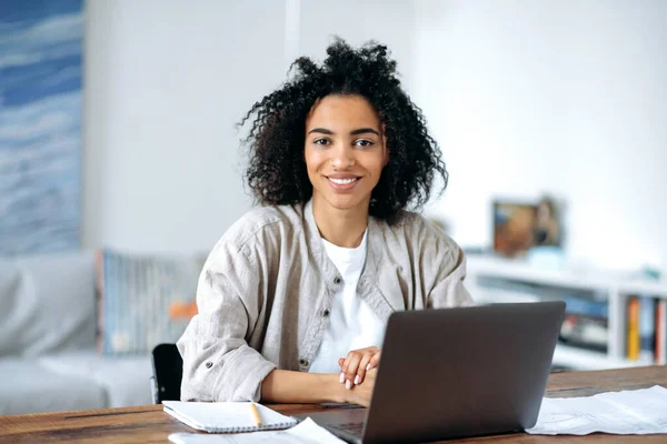 Belle amicale confiante jeune femme afro-américaine en vêtements décontractés élégants, pigiste, gestionnaire ou étudiant assis à l'ordinateur portable, travaillant ou étudiant à la maison, regardant la caméra, souriant — Photo