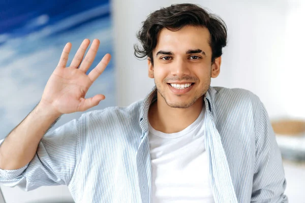 Close-up portrait of joyful successful confident young hispanic man, dressed in stylish clothes, greeting with hand gesture during online video meeting with colleagues, looks at camera, smile friendly — Stock Photo, Image