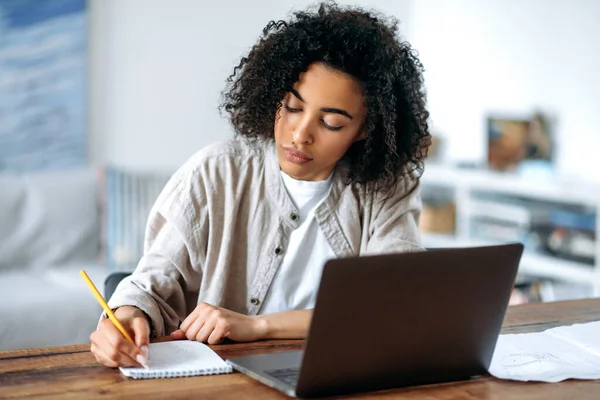 Focalisé confiant intelligent afro-américain aux cheveux bouclés fille en vêtements décontractés, pigiste ou étudiant, travailler ou étudier à distance tout en étant assis dans le salon à l'ordinateur portable et de prendre des notes — Photo