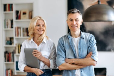 İki üst düzey mutlu beyaz erkek ve kadın meslektaşın portresi, şık giyinmiş, masanın yanında duruyor, gülümsüyor, iş kadını dizüstü bilgisayarını tutuyor, iş adamı kollarını kavuşturmuş