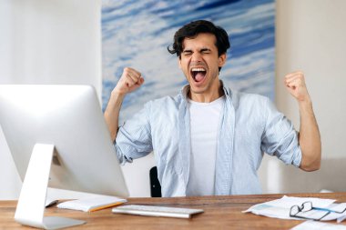 Heyecanlı, mutlu, başarılı İspanyol adam, serbest çalışan, günlük giysiler içinde, işyerinde oturuyor, zaferle, kazançla veya büyük kazançla seviniyor, elleriyle el kol hareketi yapıyor, duygusal olarak bağırıyor.