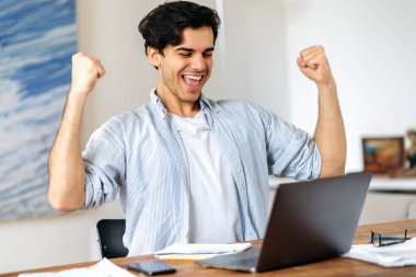 Genç, neşeli İspanyol bir adam, menajer ya da serbest çalışan, iş yerinde dizüstü bilgisayarla oturan, zaferle ya da büyük kazançla mutlu bir şekilde, anlaşma yapan, elleriyle hareket eden, mutlu bir şekilde gülümseyen.