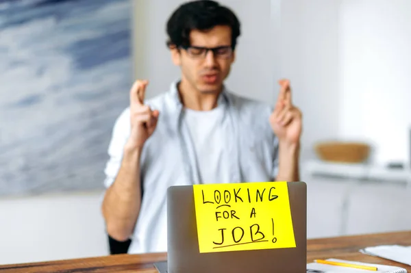 Young smart blurred hispanic man with glasses, student sits at the table, asks for support, dreams of getting a dream job crossing fingers, sign with the inscription is looking for a job on a laptop