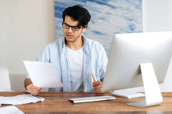 stock image Concentrated confident successful handsome hispanic man with glasses, freelancer, student or businessman working or studying rat the desk with computer, looking through business documents