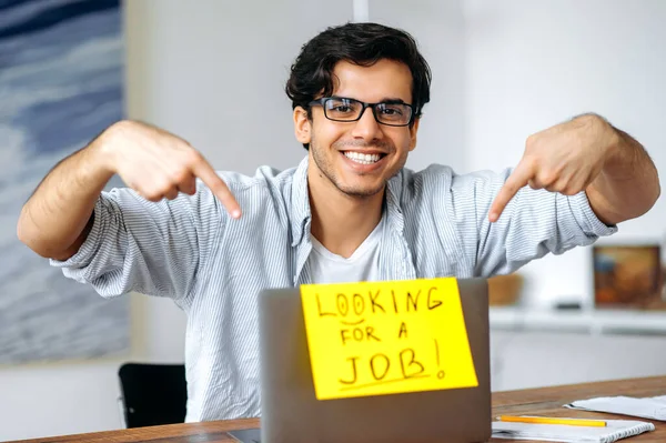 Atraente inteligente misto raça cara com óculos, estudante, sentado em sua mesa de trabalho, apontando com os dedos para um sinal pendurado em um laptop com a inscrição à procura de um emprego, olha para a câmera, sorrindo — Fotografia de Stock