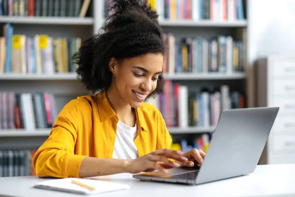 Réussie heureuse étudiante afro-américaine assise à un bureau dans une bibliothèque universitaire, avec ordinateur portable, textos avec des amis ou des camarades de classe, faire des devoirs ou se préparer à l'examen, navigation sur Internet, sourires — Photo