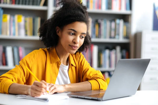 Smart terfokus indah african american perempuan siswa dalam pakaian kasual duduk di meja di perpustakaan universitas dengan laptop, menonton video pendidikan tutorial, mengambil catatan, melihat layar — Stok Foto