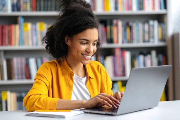 Murid perempuan afrika Amerika yang ceria dan percaya diri dengan pakaian bergaya duduk di meja perpustakaan dengan laptop, bersiap untuk ujian, SMS dengan teman-teman, browsing internet, tersenyum — Stok Foto