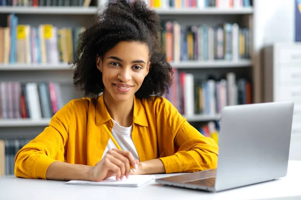 Porträtt söt framgångsrik afrikansk amerikansk kvinnlig student sitter vid bordet i universitetsbiblioteket, bär eleganta kläder, med laptop, gör läxor eller förbereda sig för examen, tittar på kameran, ler — Stockfoto
