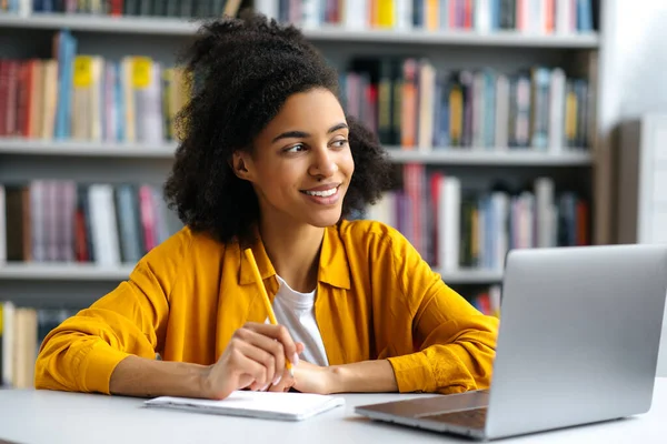 Joyeux succès jolie frisée étudiante afro-américaine aux cheveux assis à table dans la bibliothèque du campus en vêtements décontractés, avec ordinateur portable, faire des devoirs ou se préparer à l'examen, détourner les yeux, sourire — Photo