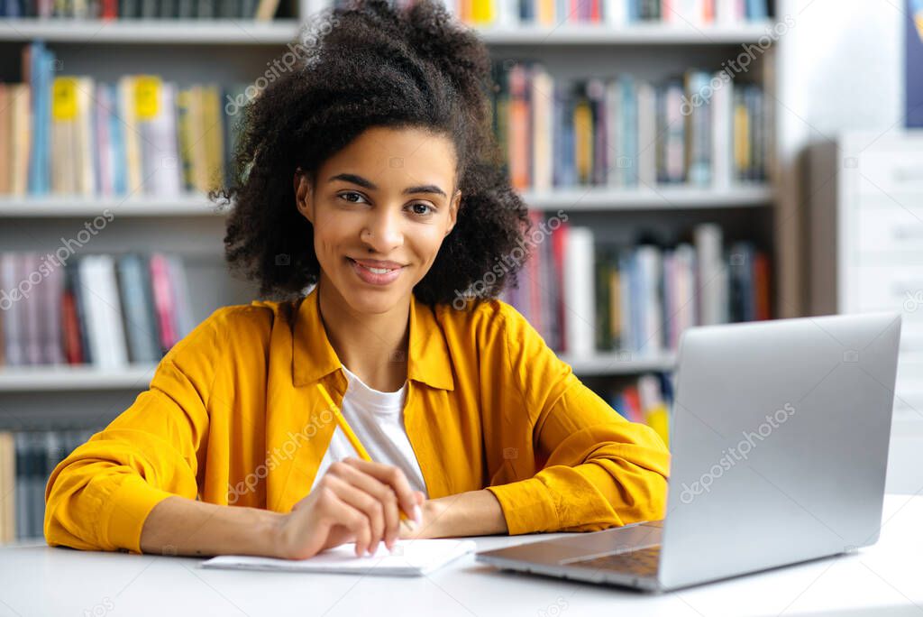 Portrait cute successful african american female student sitting at table in university library, wearing stylish clothes, with laptop, doing homework or preparing for exam, looking at camera, smiling