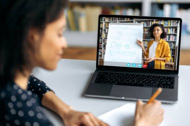 Bir bayan öğretmenle dizüstü bilgisayar ekranında Afro-Amerikalı zeki bir bayan öğrencinin omzunun üstünden bakın. Kadın öğretmen çevrimiçi ders veriyor, kız öğrenci not alıyor. Çevrimiçi eğitim, video çağrısı