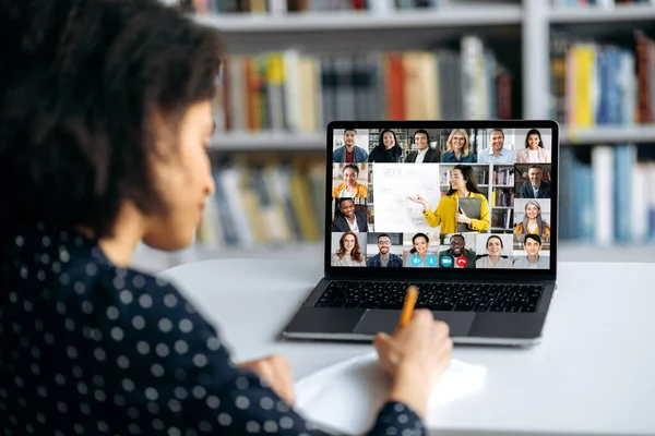 View over shoulder of an mixed race smart female student at a laptop screen with a female teacher and students. Преподаватель ведет онлайн лекцию, студентка делает заметки. Онлайн тренинг, видеозвонок — стоковое фото