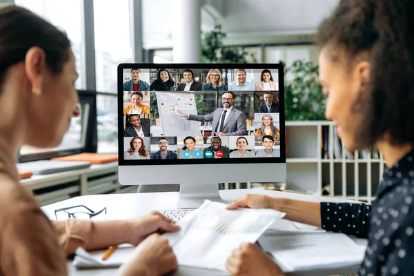 Online επαγγελματική συνάντηση, καταιγίδες ιδεών. Προβολή πάνω από τους ώμους δύο γυναικών σε μια οθόνη υπολογιστή με διαφορετικούς επιτυχημένους πολυφυλετικούς ανθρώπους που συγκεντρώθηκαν σε ένα βίντεο συνέδριο για να συζητήσουν θέματα εργασίας — Φωτογραφία Αρχείου