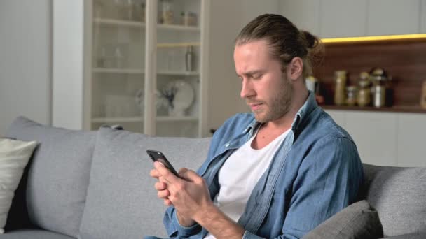 Chico americano muy alegre en una camisa de mezclilla elegante se sienta en el sofá en la sala de estar, utiliza un teléfono inteligente, navegar por Internet, mensajes de texto con amigos, se regocija para ganar o una buena noticia, sonriendo felizmente — Vídeo de stock