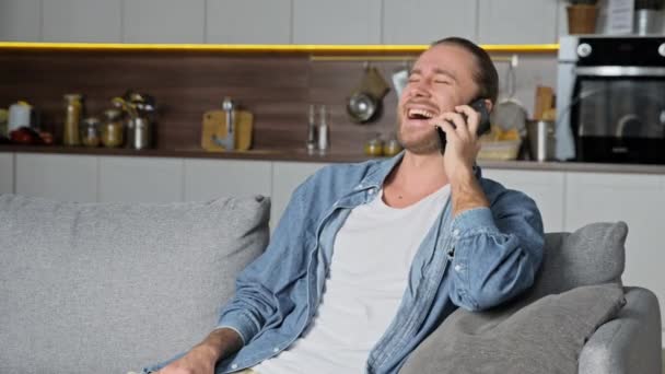 Smartphonegesprek. Blanke vrolijke man in stijlvolle casual kleding, zitten op de bank in de woonkamer, een aangenaam telefoongesprek met een vriend of vriendin, glimlachen — Stockvideo