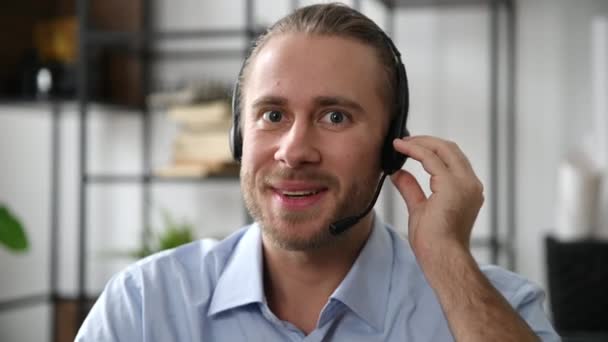 Close-up de um operador amigável barbudo atraente de call center, especialista, trabalhador de serviço de suporte ou líder de negócios. Homem caucasiano em fone de ouvido realiza consulta on-line, olha para a câmera, sorrindo — Vídeo de Stock