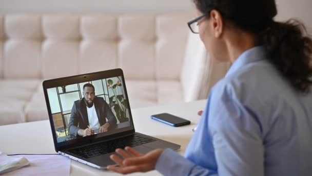 Reunião de negócios online via videoconferência. Vista sobre o ombro de uma mulher de negócios para uma tela de laptop com colega afro-americano, colegas de trabalho conversando usando um aplicativo de comunicação on-line — Vídeo de Stock