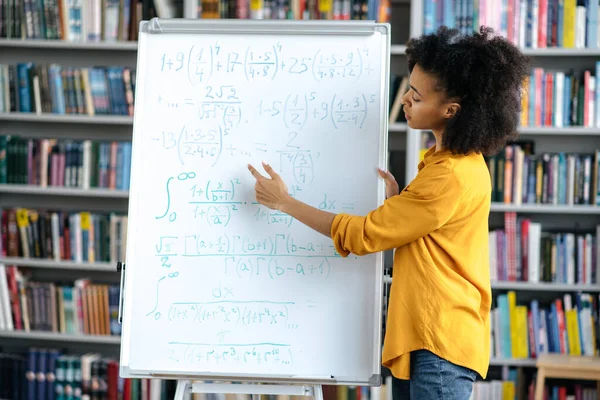 Confident African American intelligent young adult female teacher stand near the whiteboard, conducts lecture at the library, shows information to the students. Distant learning, online conference