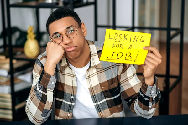 Upset unemployed young clever mixed race man with glasses, student or freelancer, holds a sign with the inscription looking for a job, hopes and waits to get a dream job, sadly looks at camera — Stock Photo, Image