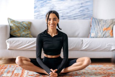 Afro-Amerikan spor kıyafetleri içinde neşeli, hoş Afro-Amerikan genç bir kadın, evde lotus pozisyonunda halının üzerinde oturuyor, yoga yapıyor, esniyor, sağlıklı bir yaşam sürüyor, kameraya bakıyor, gülümsüyor.