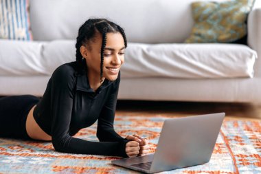 Güzel vücutlu, spor giyimli mutlu Afrikalı genç bir kadın, evde yerde yatıyor, dizüstü bilgisayar kullanıyor, video fitness veya yoga dersleri izliyor, spor yapmaya gidiyor, sağlıklı bir hayat sürüyor, gülümsüyor.